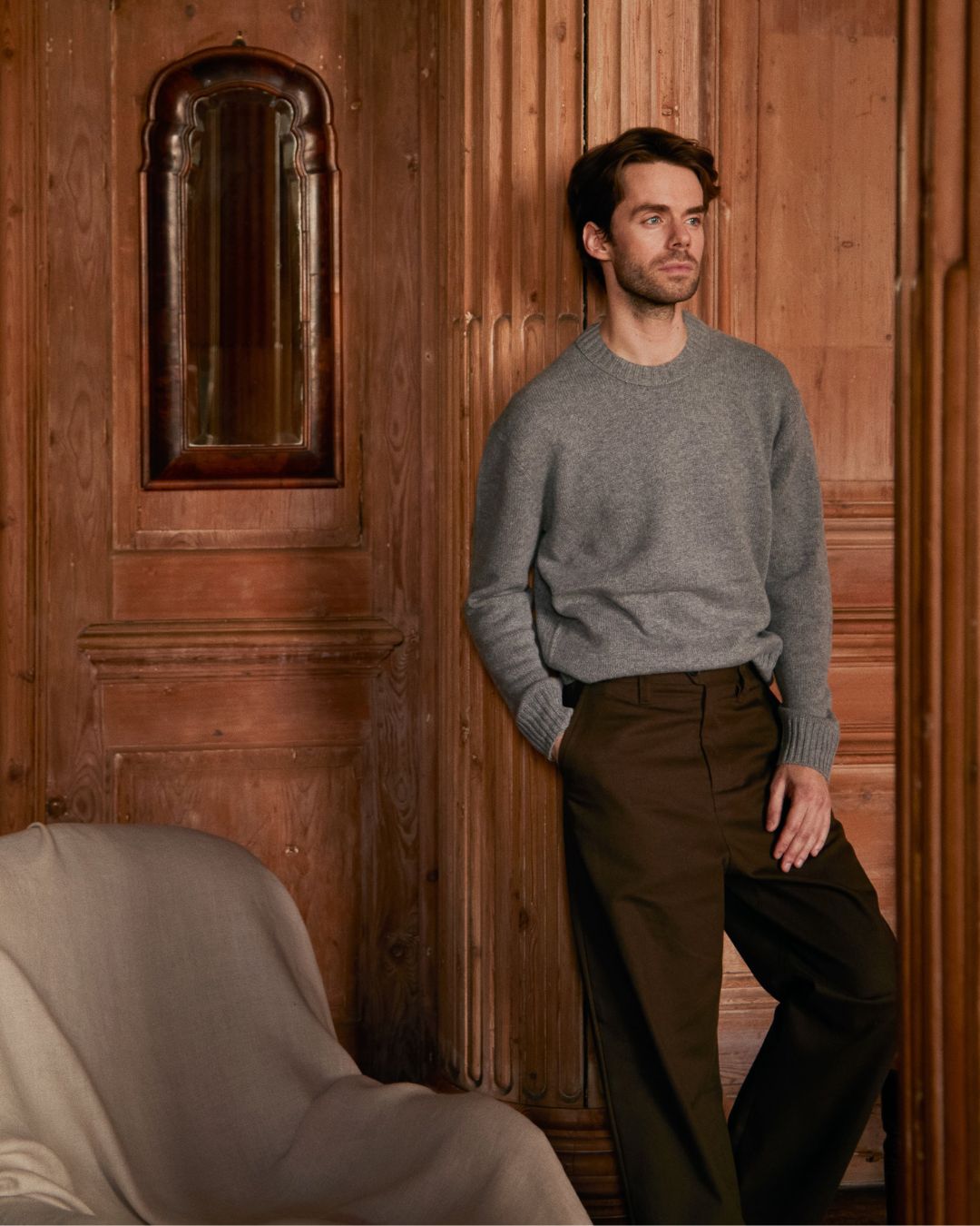 Man leaning against carved warm wood paneling wearing a grey wool crewneck
