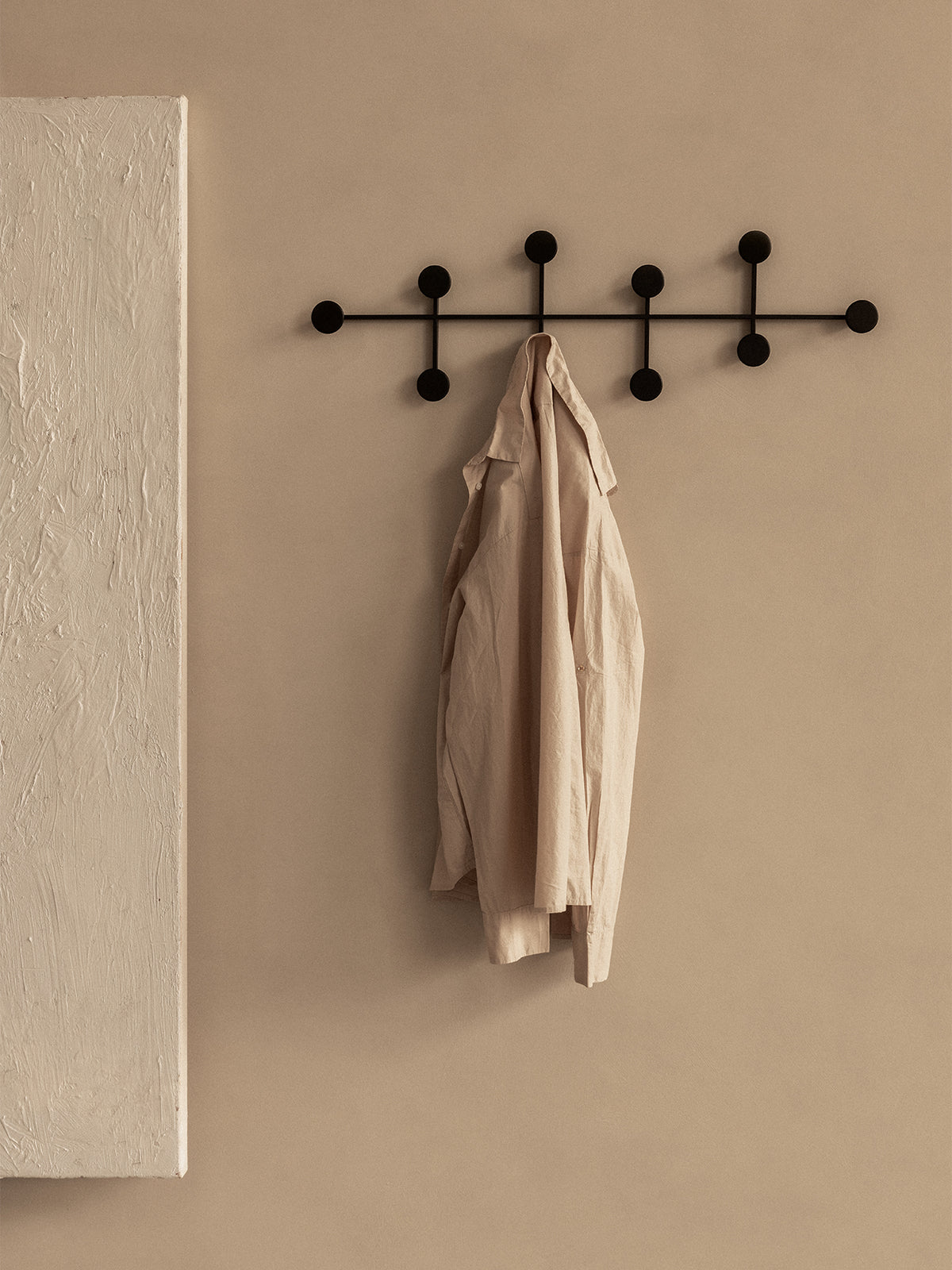 Schwarzer Afteroom Coat Hanger Large an beiger Wand mit einem beigen Leinenhemd.