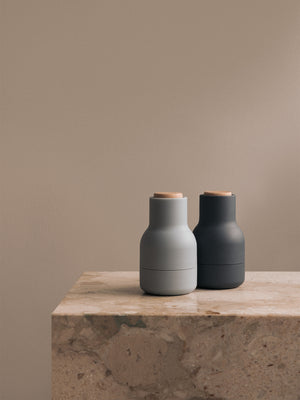 Two Audo Bottle Grinders, one light grey and one dark grey, standing on the corner of a beige marble plinth against a wall.