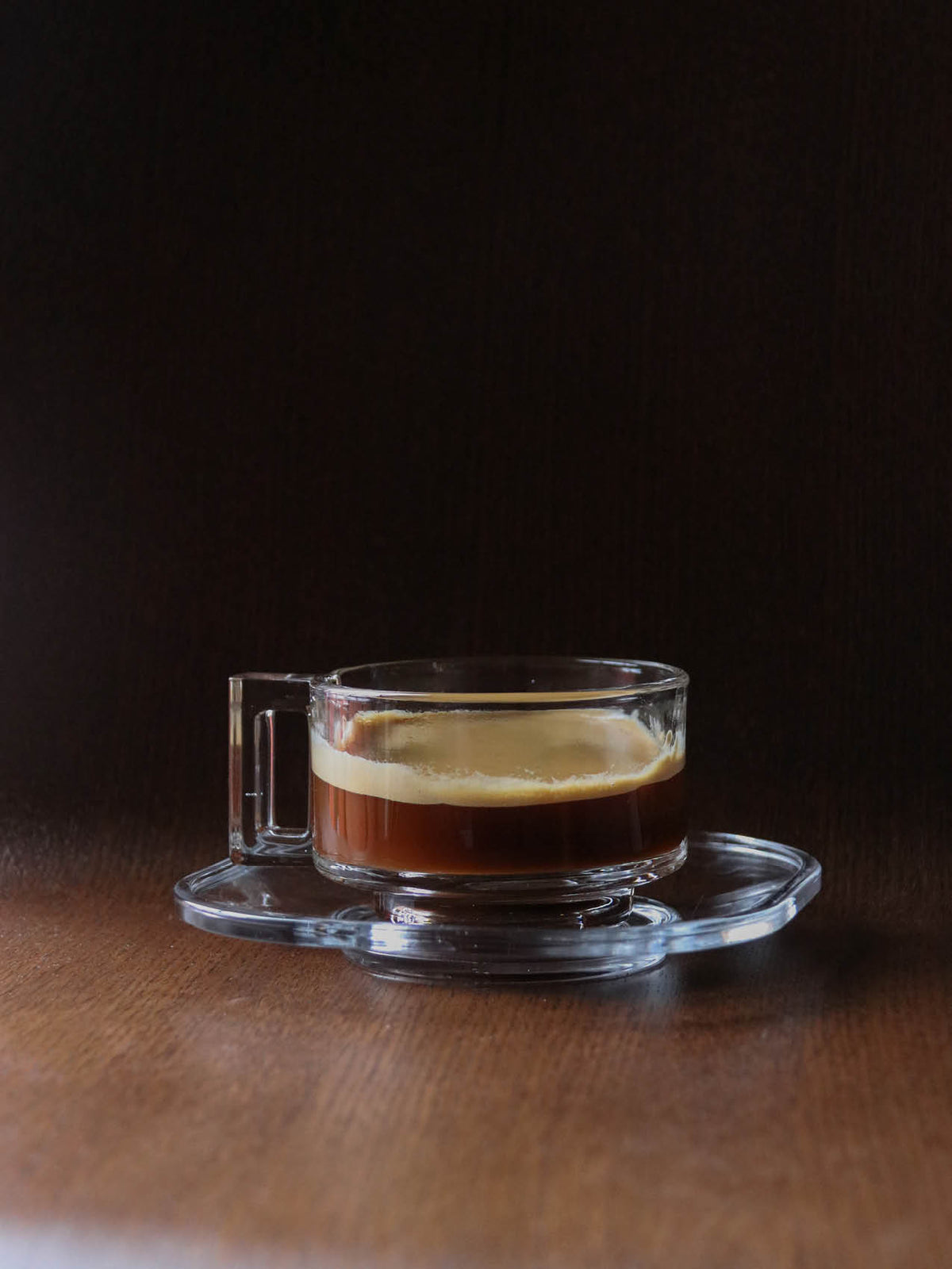 Clear double-walled glass espresso cup and saucer on wooden table, dark crema