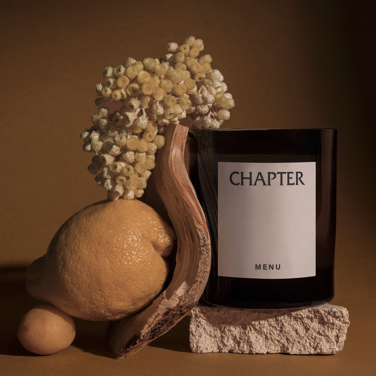 Dark brown glass candle labeled "CHAPTER MENU" beside driftwood, pale yellow dri