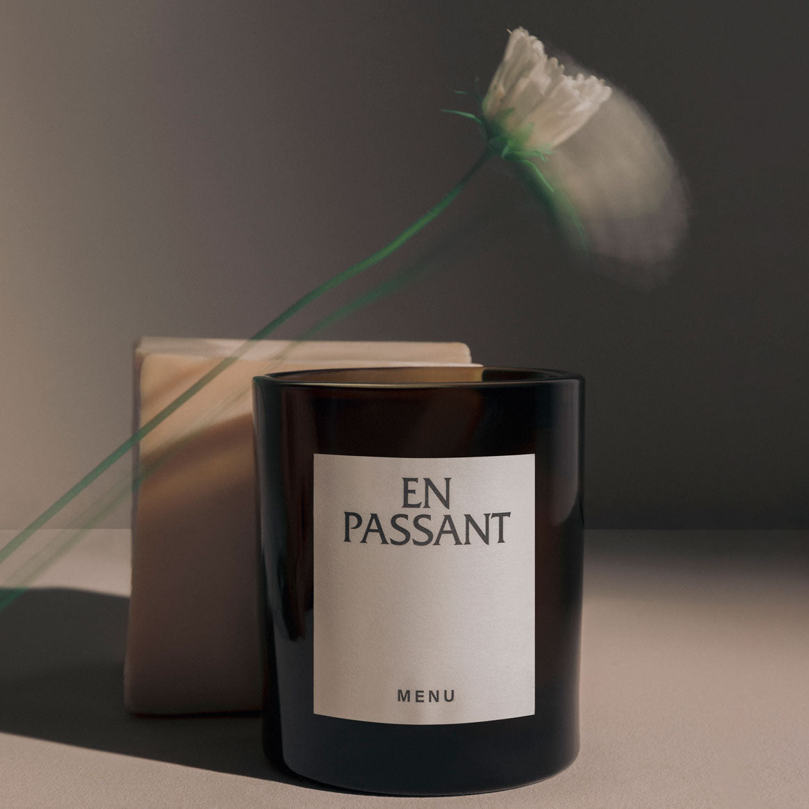 Dark brown glass candle with white "EN PASSANT" label, blurred white flower