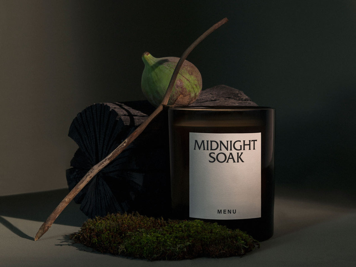 Dark glass candle labeled "MIDNIGHT SOAK" beside green fig, twig, black folded