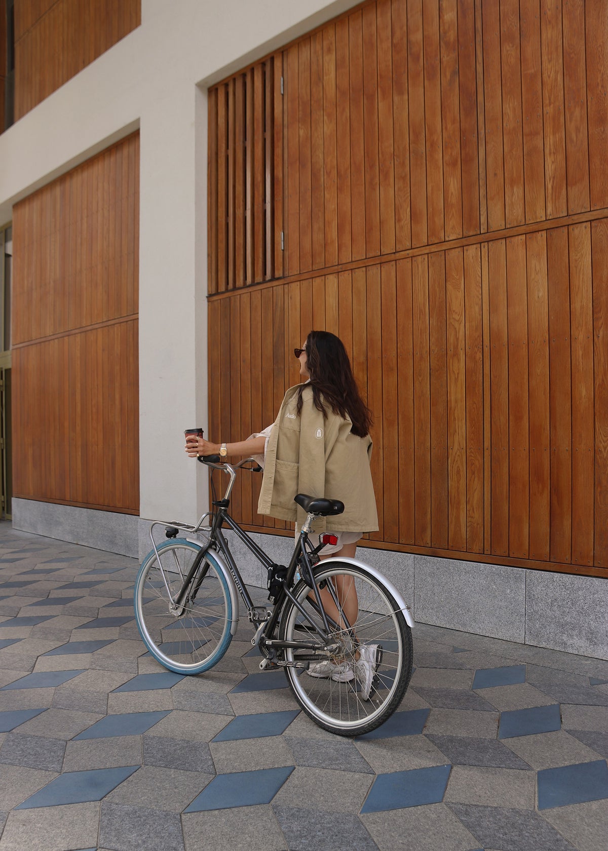 Woman in beige jacket holding coffee beside black city bike against tall