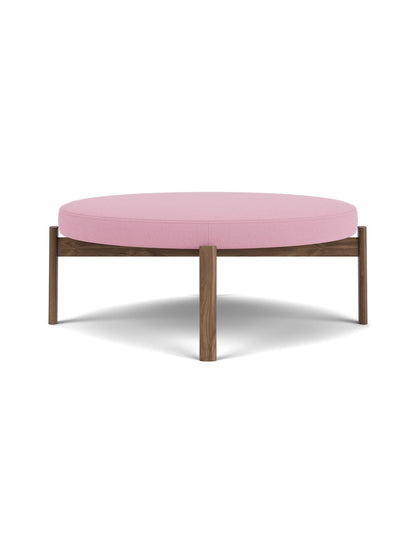 A front view of the circular Passage Pouf, showing its thick pink upholstered cushion on a dark brown wooden frame and legs.