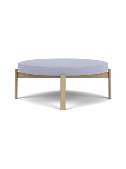 A front-facing view of the Passage Pouf, featuring a circular, light purple upholstered seat and a natural oak wooden frame.