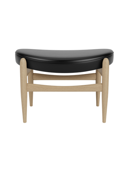 Elizabeth Ottoman featuring a curved black leather seat and a light natural oak frame with tapered legs.