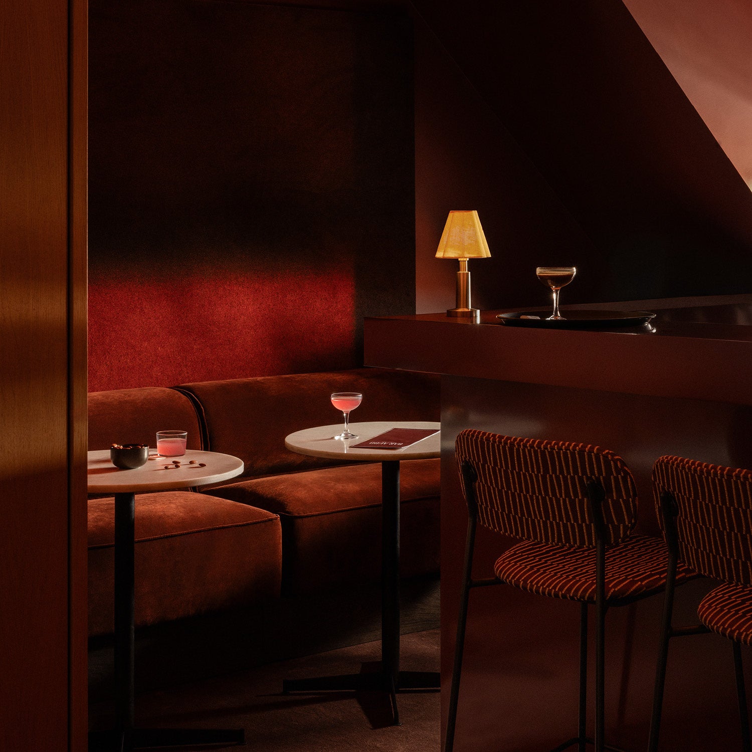 Cozy bar nook with curved rust velvet banquette, round white marble tables, striped orange bar stools and amber table lamp