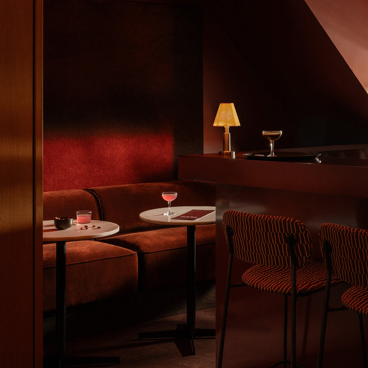 Cozy bar nook with curved rust velvet banquette, round white marble tables, striped orange bar stools and amber table lamp