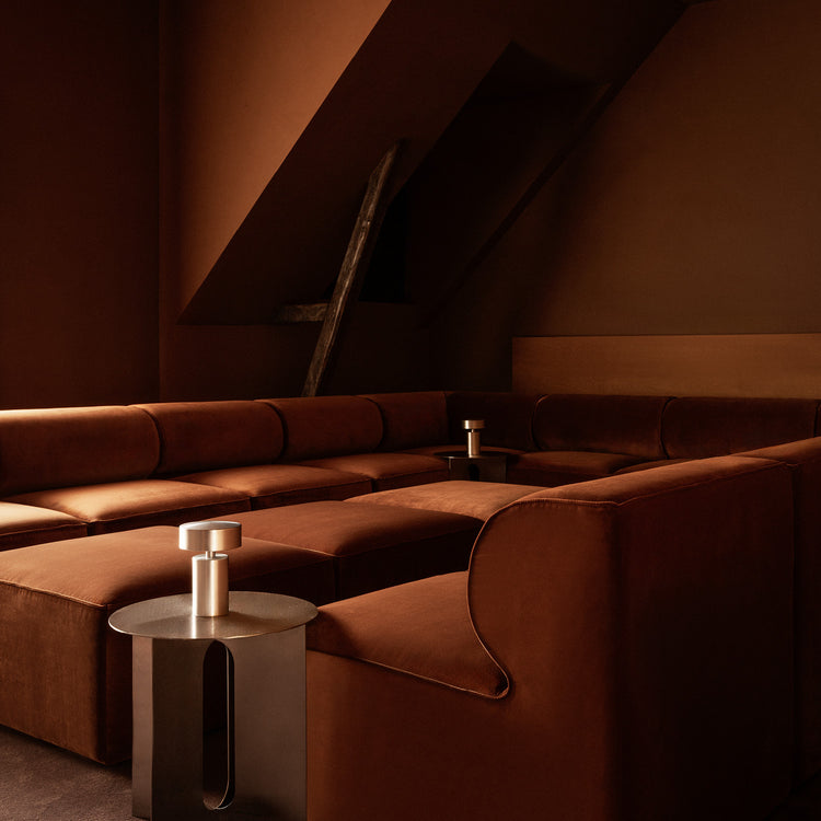 Deep rust-brown velvet modular sofa in corner layout with curved armrest, metal side tables and warm matte walls