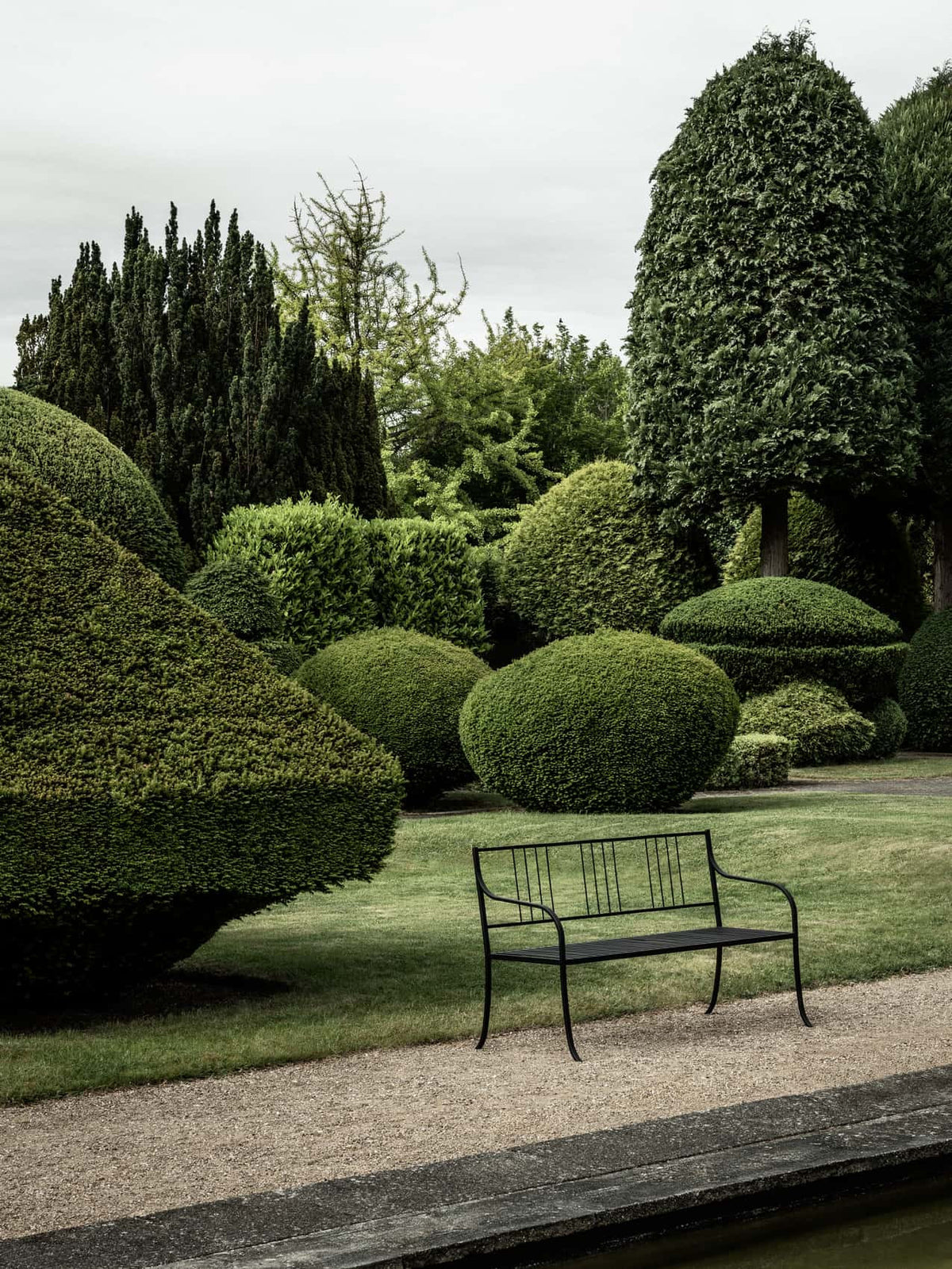 Bloss Garden Bench in black powder-coated steel placed on a gravel path in a lush green garden with sculpted topiary trees.