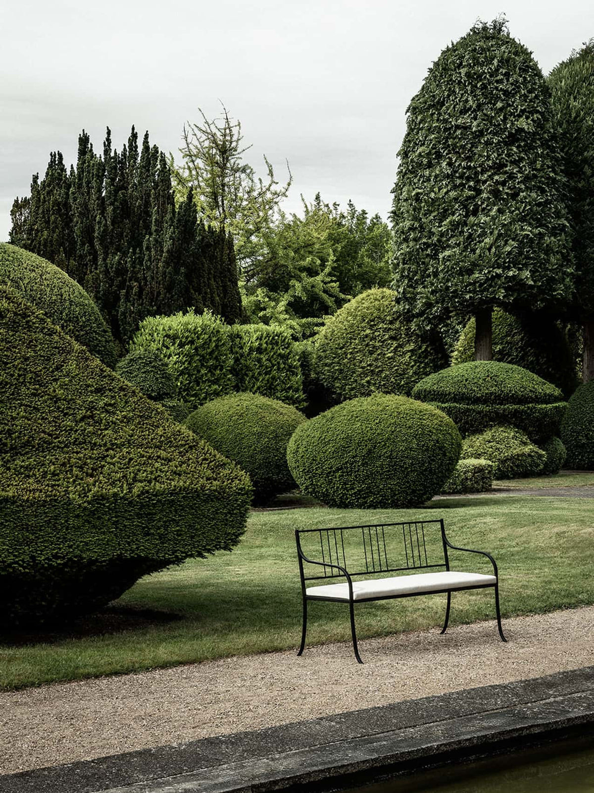 Black powder-coated steel Bloss Garden Bench with a cushion set, placed in a lush garden with sculpted hedges.