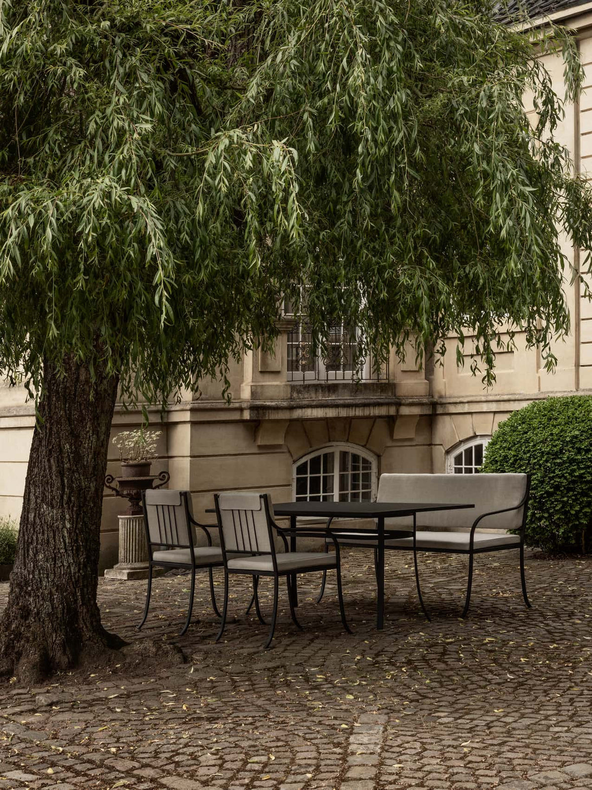 Bloss Garden Dining Chair, Table and Bench set in black powder-coated steel on a terrace beneath a weeping willow tree.