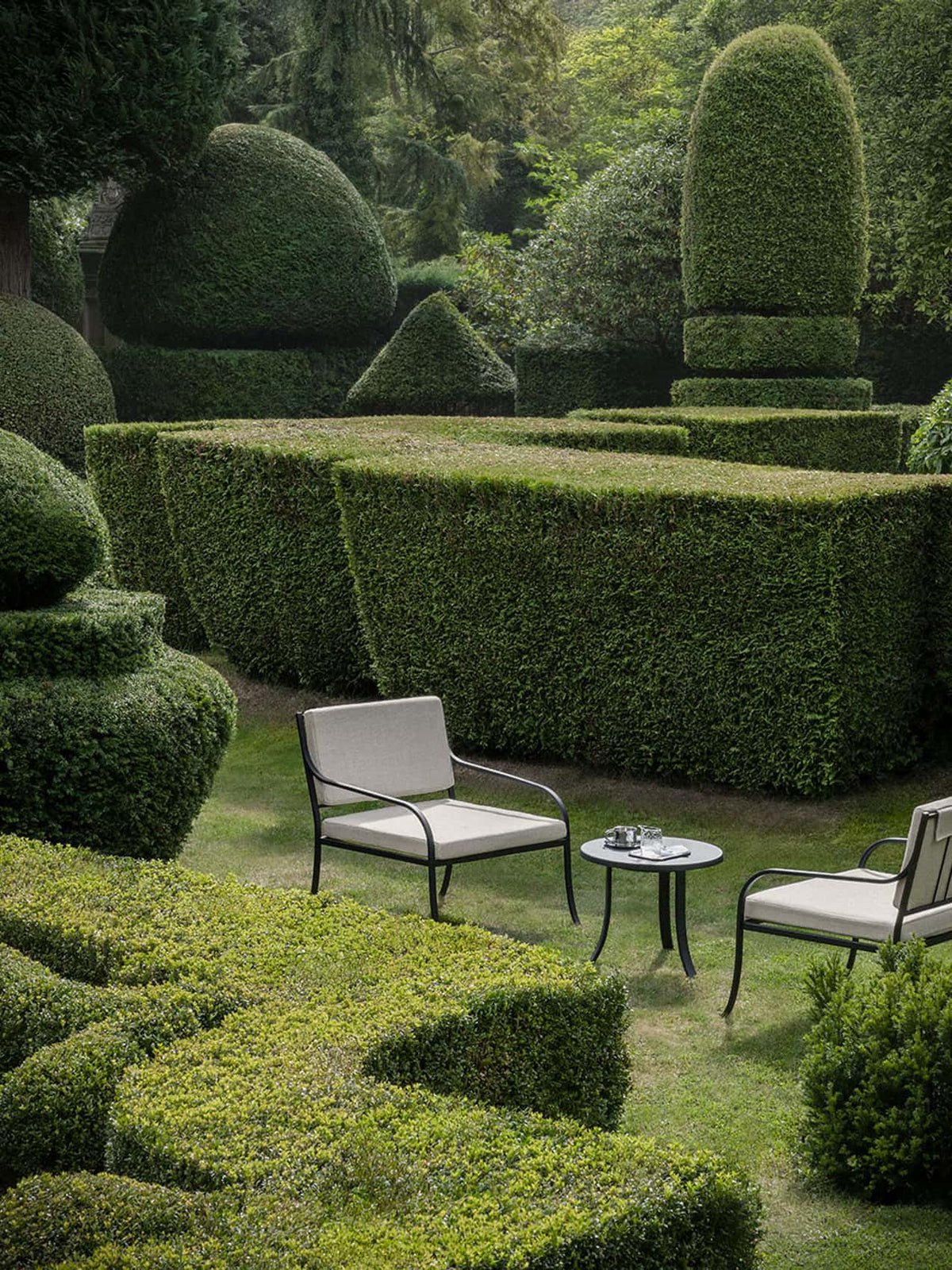 Bloss Garden Side Table and lounge chairs in black powder-coated steel with cushions, placed outdoors amidst topiary hedges.