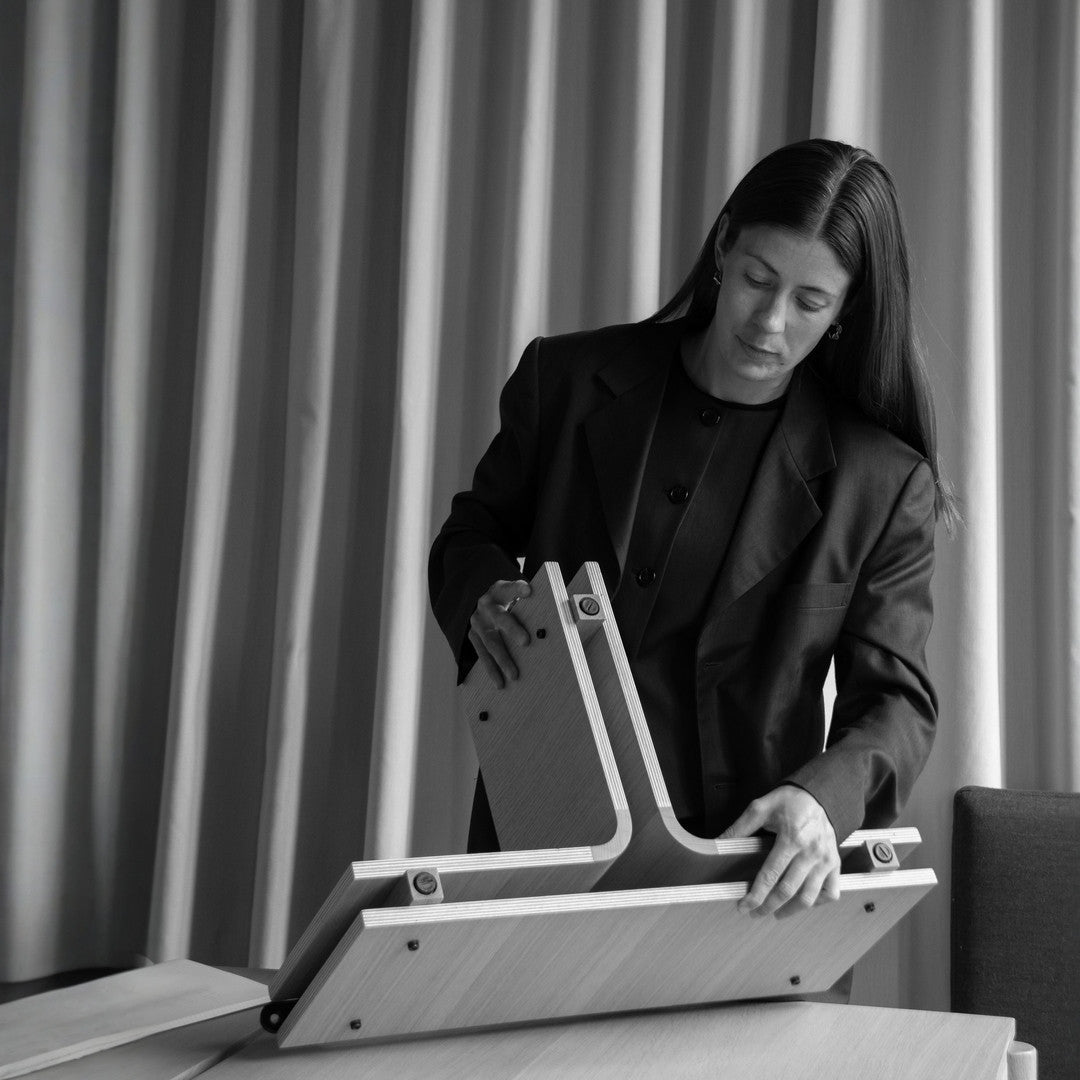 Laura Lange assembling wooden furniture components in a minimalist interior setting with draped curtains.