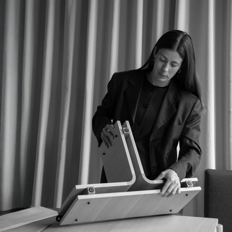 Laura Lange assembling wooden furniture components in a minimalist interior setting with draped curtains.