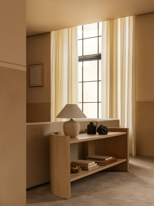 A light oak Crescent Shelving unit behind a beige Eave Modular Sofa, styled with a Torso Table Lamp and books on a Duomo Rug.