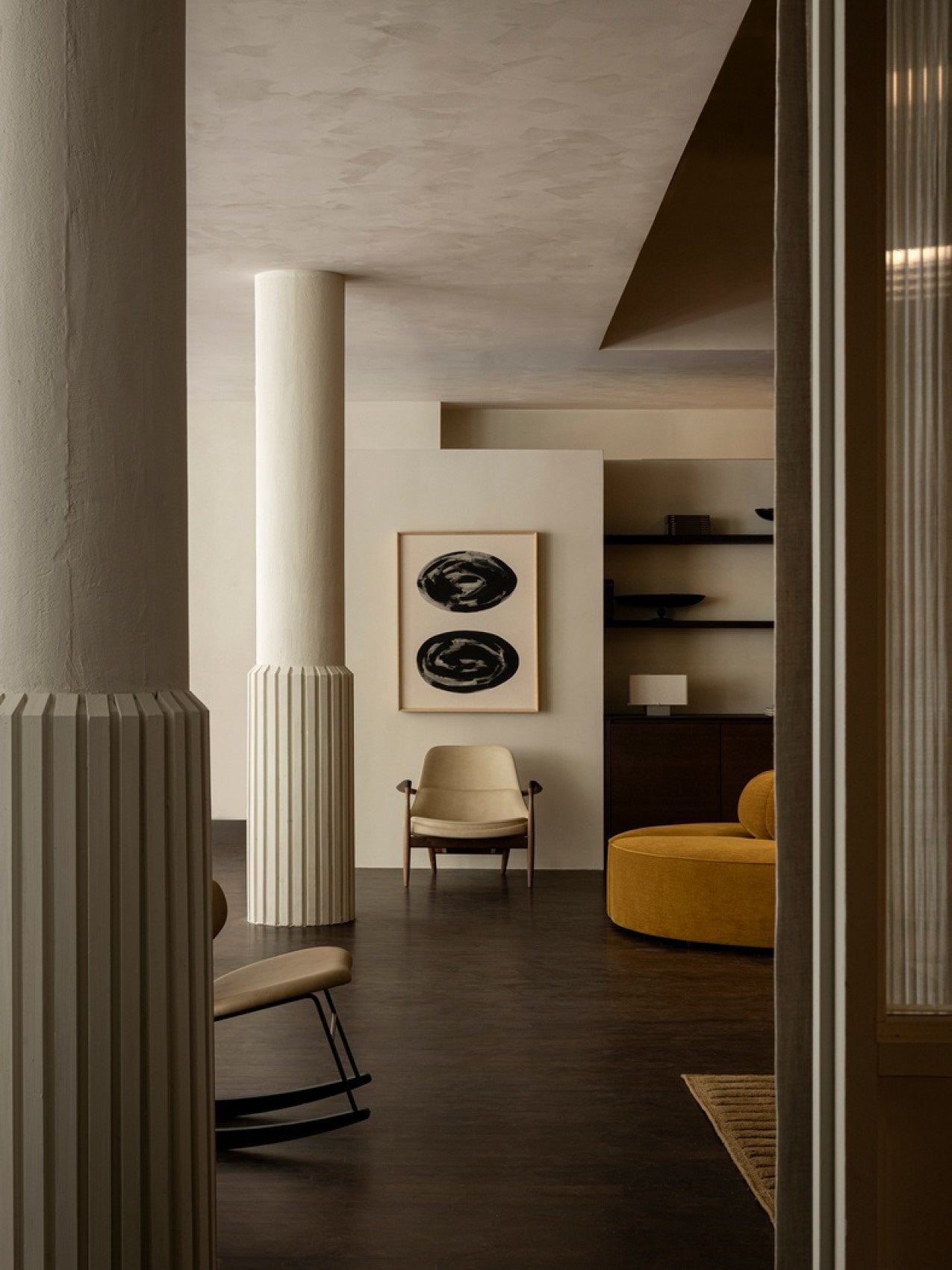 Minimal living room with beige leather armchair, fluted cream columns, dark wood floor and mustard round sofa.