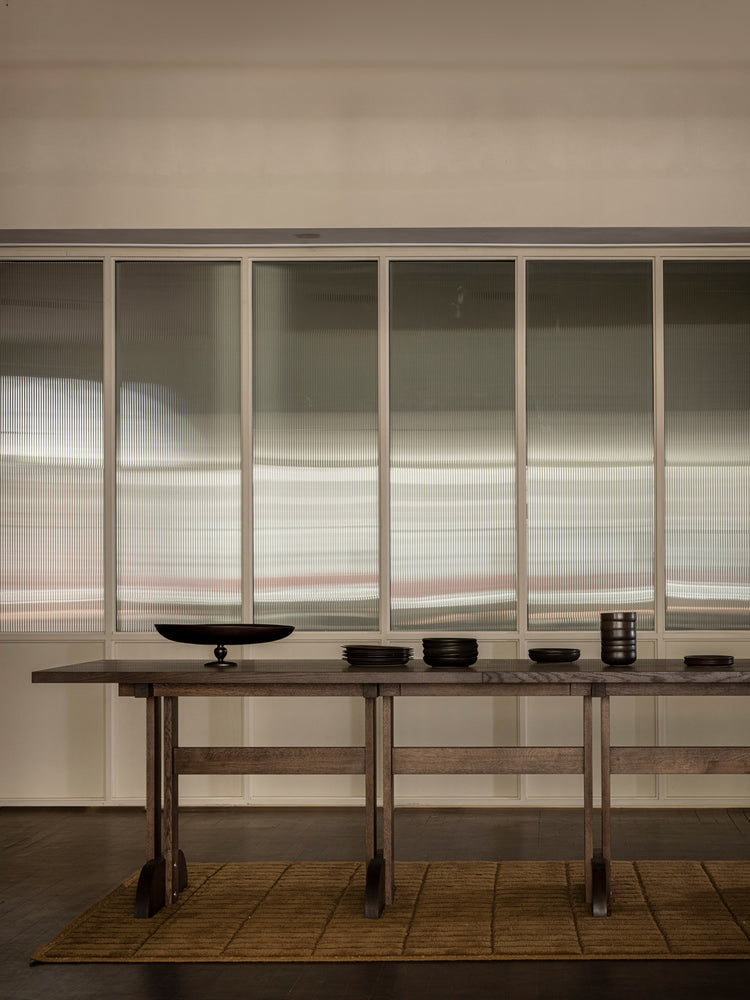 Long wooden console table with dark wood bowls and stacked ceramic cups in front of frosted glass paneled wall.