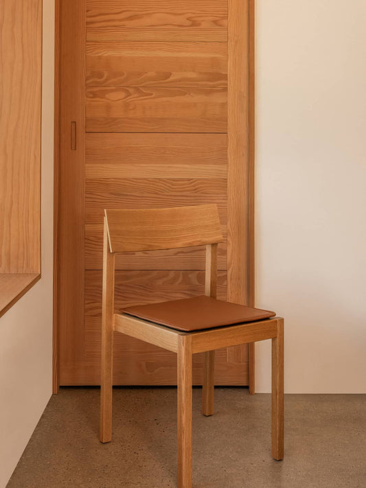 Mauro Dining Chair in natural oak with a brown leather seat cushion, positioned against a wooden door and light wall.