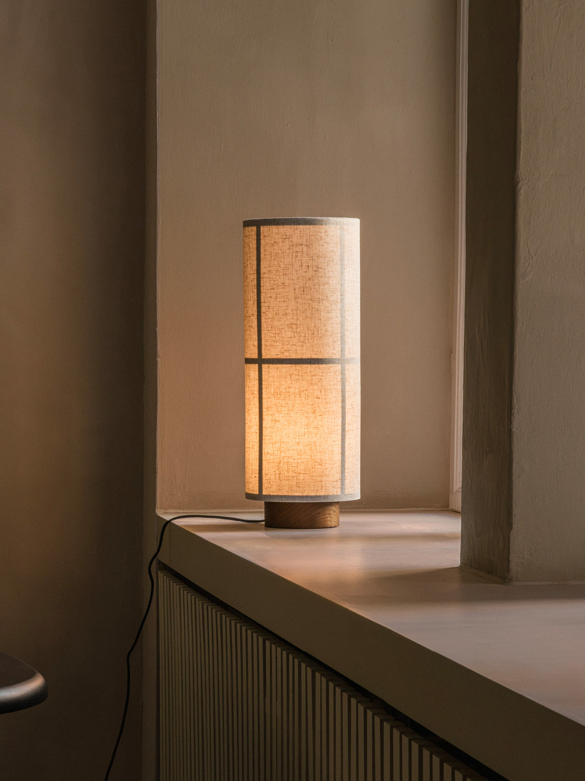 Hashira Table Lamp in raw linen standing on a windowsill, glowing softly and diffusing warm light.