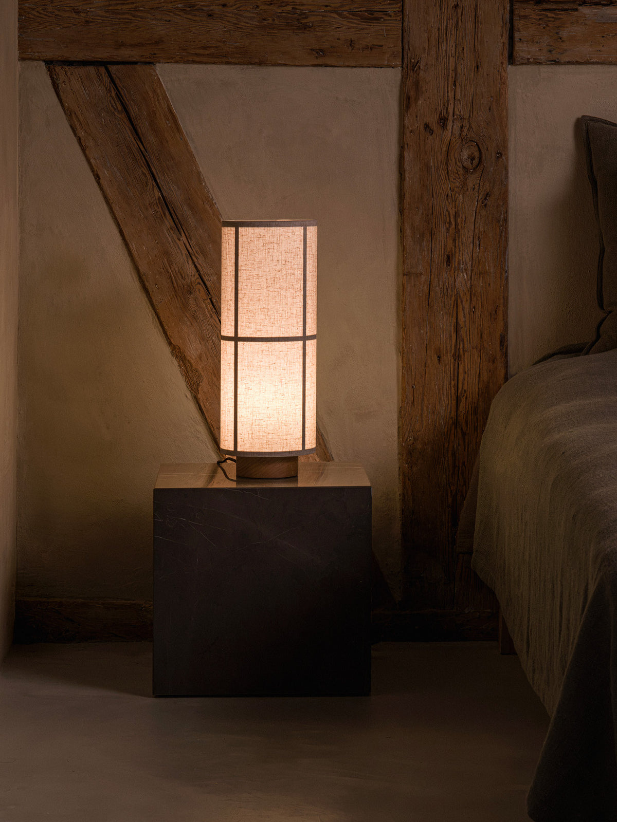 Hashira Table Lamp in raw linen, placed on a cubic dark plinth next to a bed in a rustic interior setting.