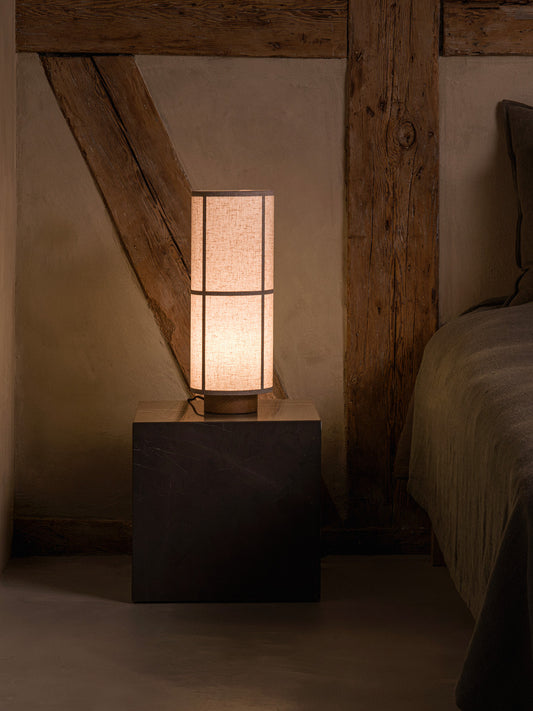 Hashira Table Lamp in raw linen, placed on a cubic dark plinth next to a bed in a rustic interior setting.