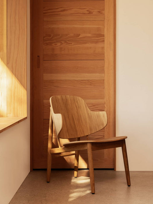 The Penguin Wooden Lounge Chair in natural oak, positioned in a sunlit room against a matching wooden door.