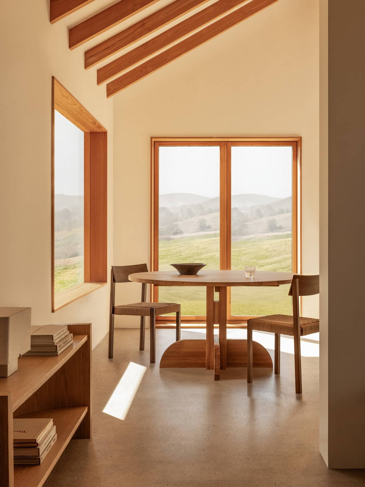 Oak Puffin Dining Table and Mauro Dining Chairs in a sunlit room with Crescent Shelving and views of rolling green hills.