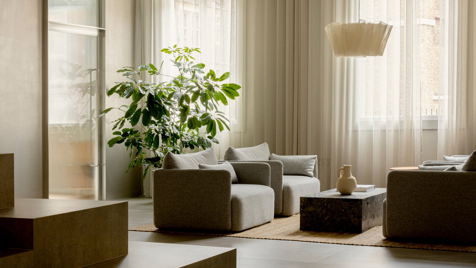 Minimal living room with gray upholstered cube armchairs, marble cube coffee table, beige pendant and large green plant.