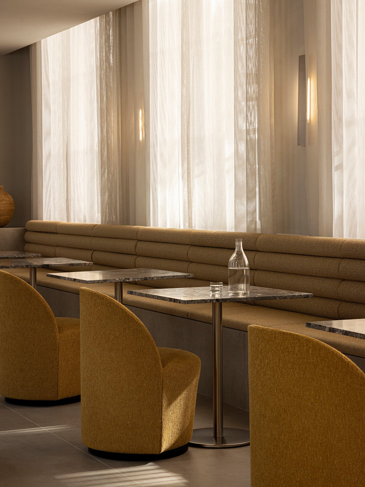 Modern cafe seating with mustard boucle upholstered chairs and banquette, marble-topped tables and sheer cream curtains.