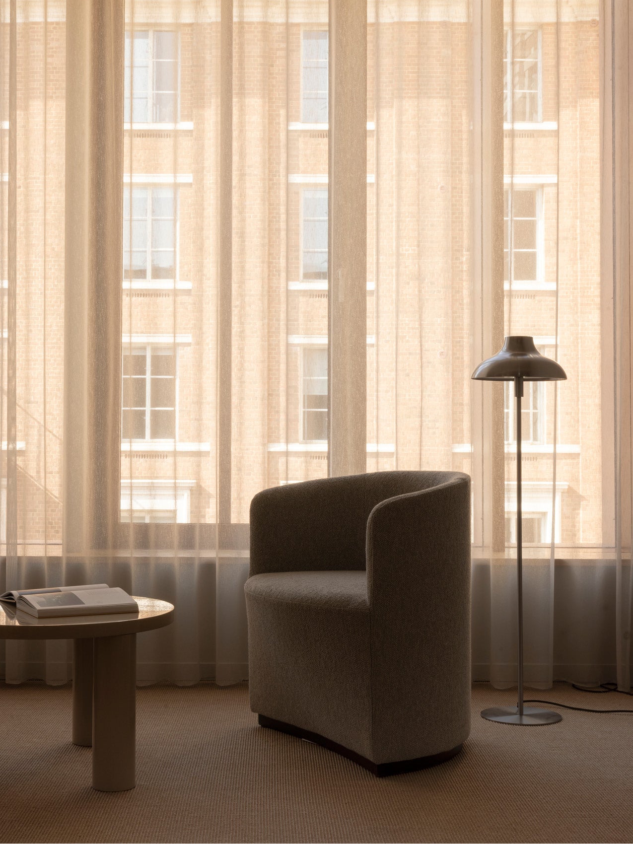 Curved beige upholstered lounge chair beside a round wood side table and slim metal floor lamp in front of sheer beige