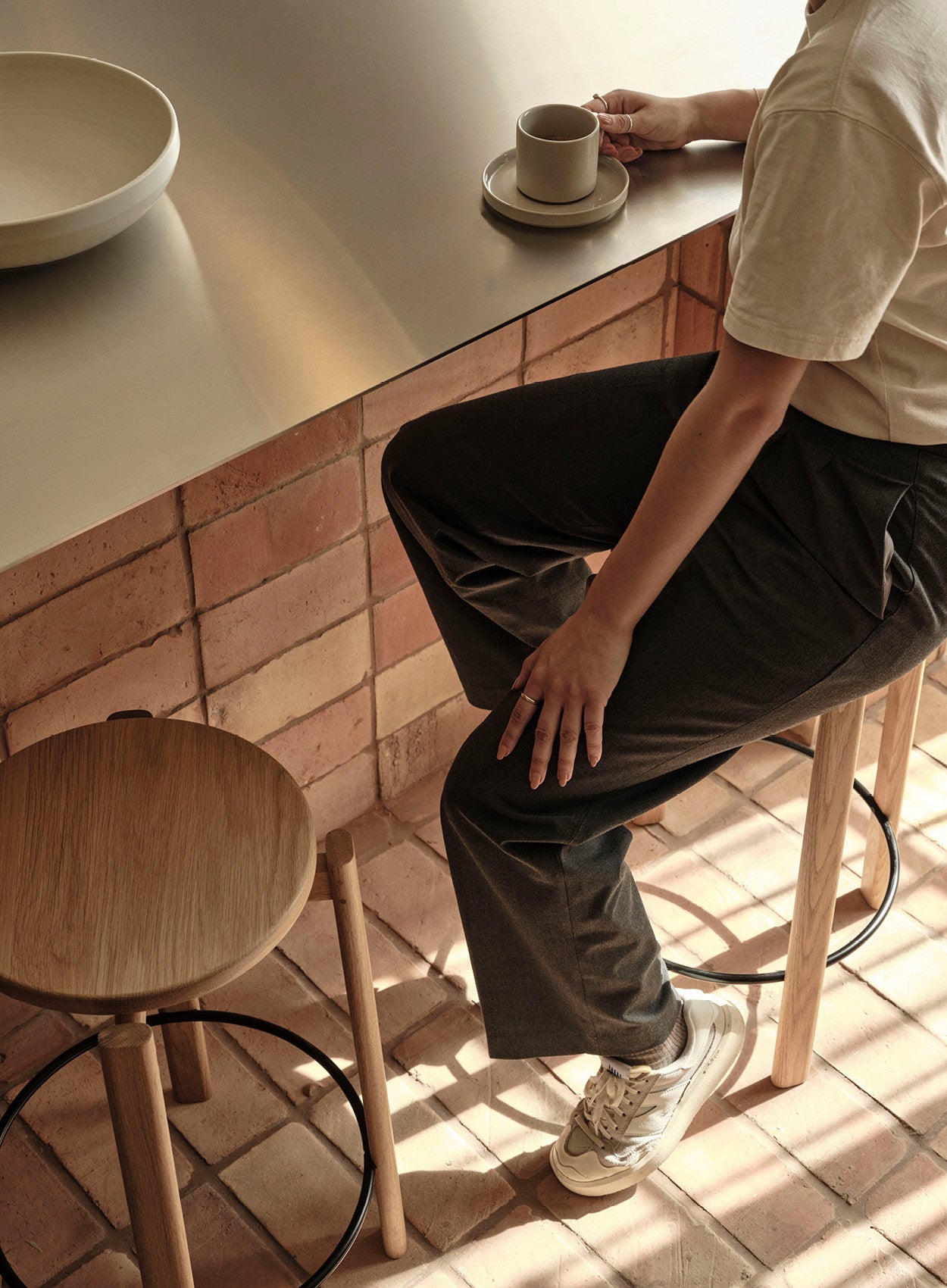 Minimal beige ceramic cup and saucer on a matte metal counter, person seated on oak bar stool beside terracotta tile wall.