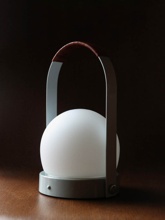 Carrie Portable Lamp with a leather handle and white opal glass shade, set on a wooden surface in a dark room.