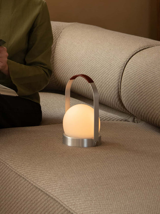 A glowing Carrie Portable Lamp with a brushed aluminium frame and leather handle rests on a beige textured sofa.