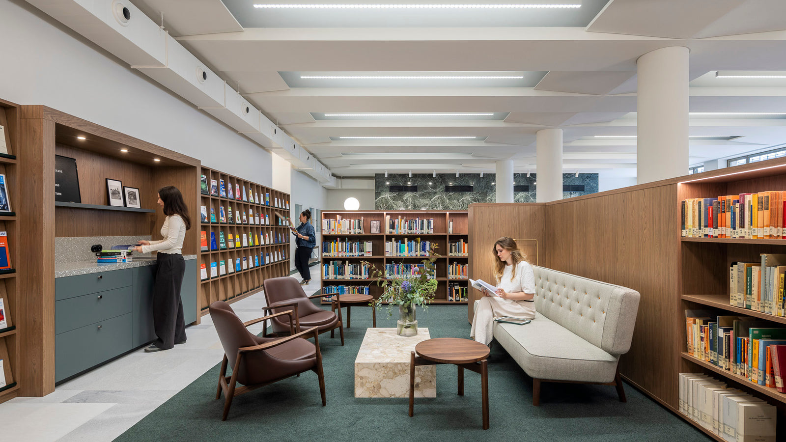 Modern library lounge with light grey tufted sofa, brown leather chairs, marble coffee table, wood shelves and people reading