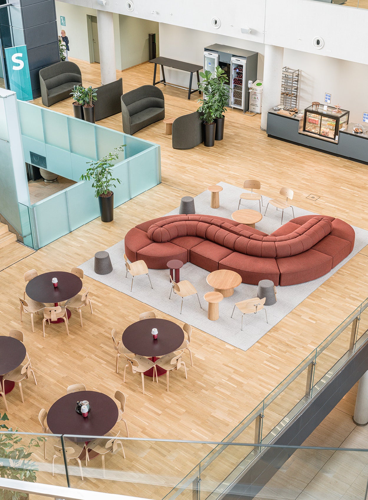 Open-plan atrium with curved rust-red modular sofa, round wooden tables, pale wood chairs and light parquet flooring