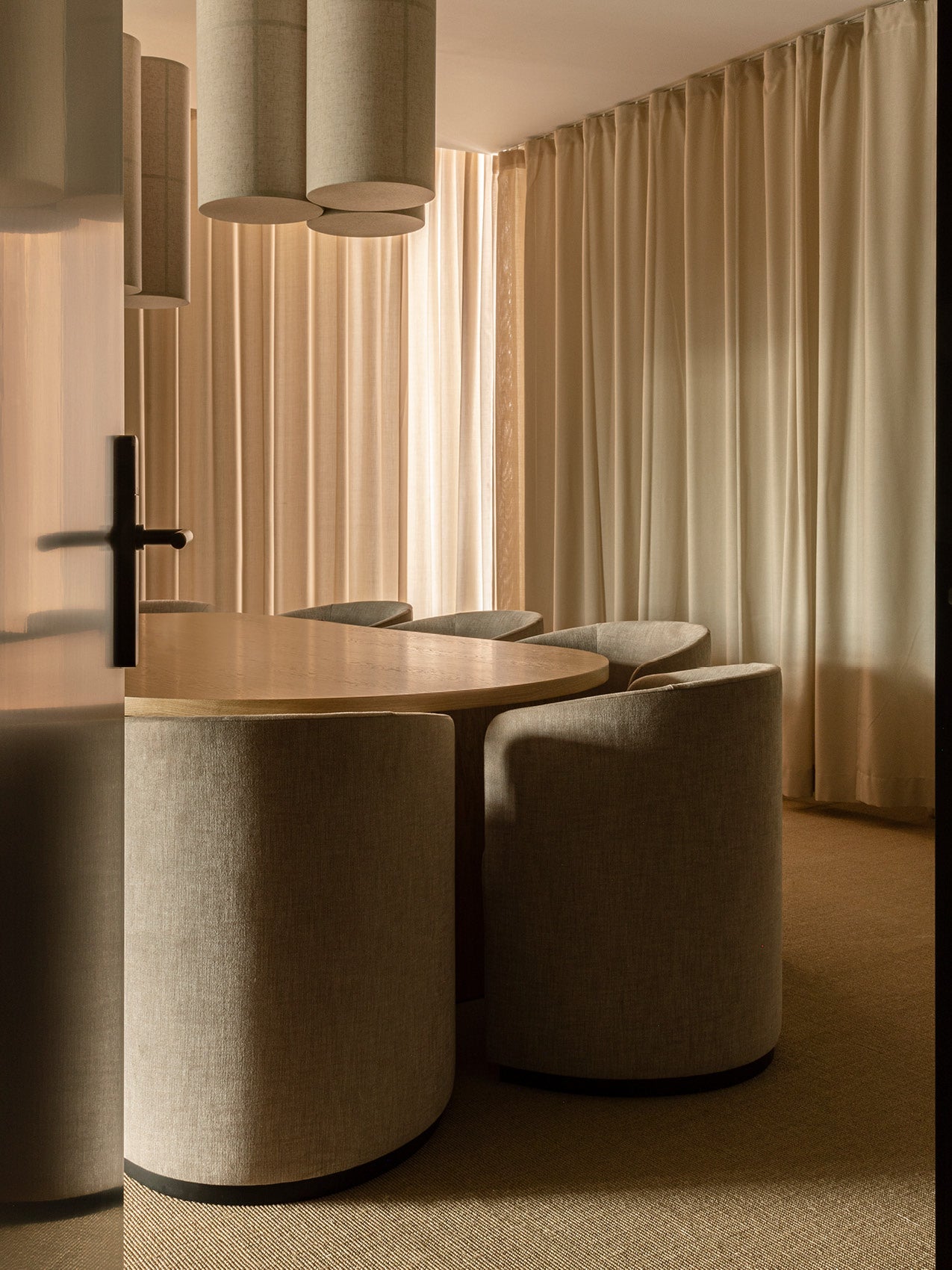 Curved oak dining table with upholstered beige barrel chairs under cylindrical fabric pendant lights, soft beige curtains