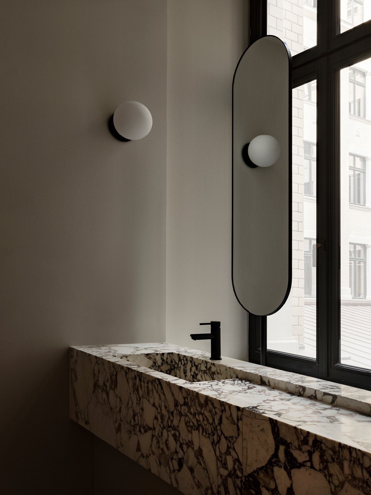 Marble double sink with dark veining, matte black faucet, oval vertical mirror and round frosted wall sconce by window