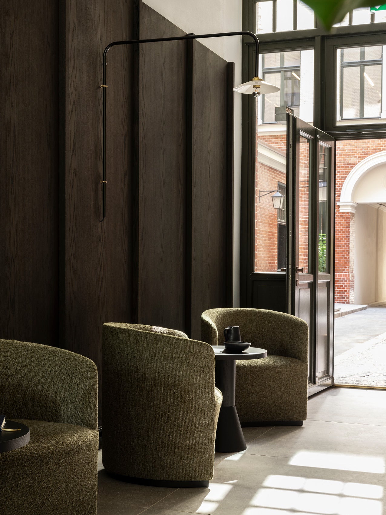 Pair of olive-green upholstered barrel chairs with black side table and matte black wall lamp by open glass door