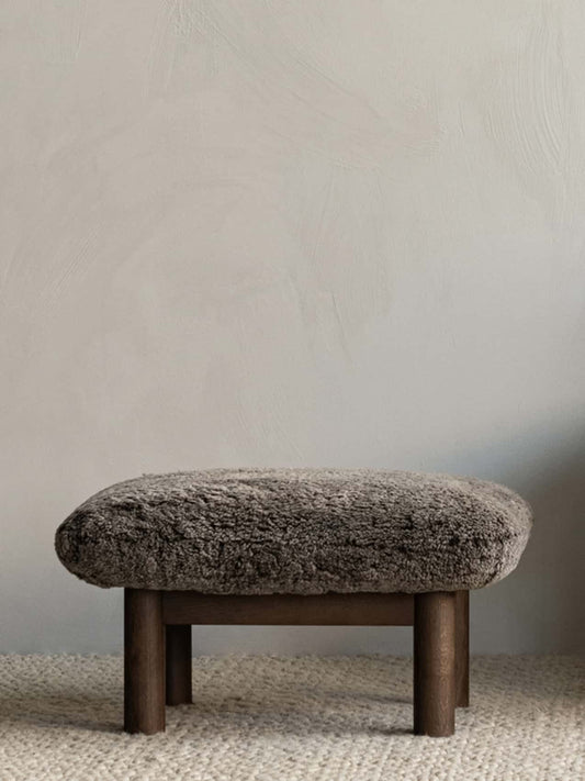 Brasilia Ottoman upholstered in soft sheepskin with robust wooden legs, placed against a neutral textured wall.