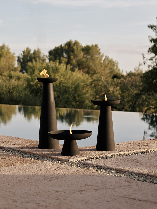 Three matte black cone-shaped outdoor fire bowls of varying heights with small flames by a reflective pool and trees.