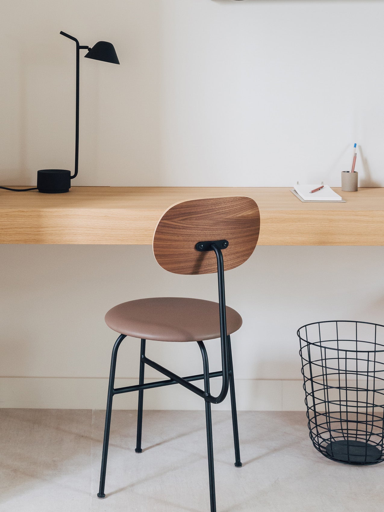 Minimal wooden desk with black metal-legged stool, brown seat and curved walnut backrest, black desk lamp and wire