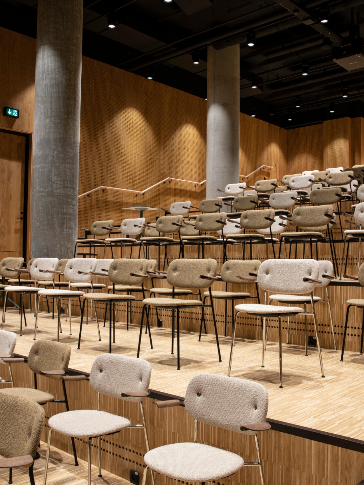 Tiered auditorium with beige wool-upholstered chairs, rounded backrests, wooden armrests and slim black metal legs.