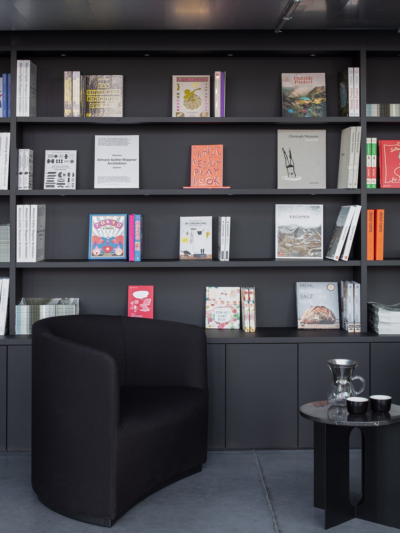 Black upholstered curved armchair beside black side table and dark grey built-in bookshelves displaying colorful books.