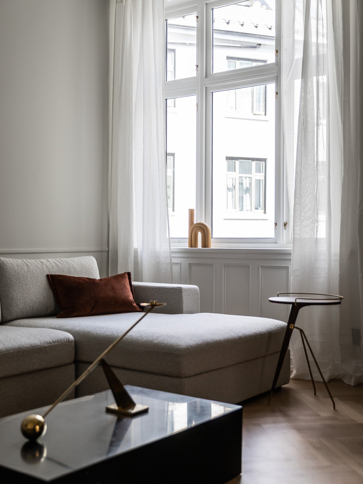 Minimal living room with light grey upholstered chaise, rust-brown cushion, marble coffee table and arched wooden sculpture