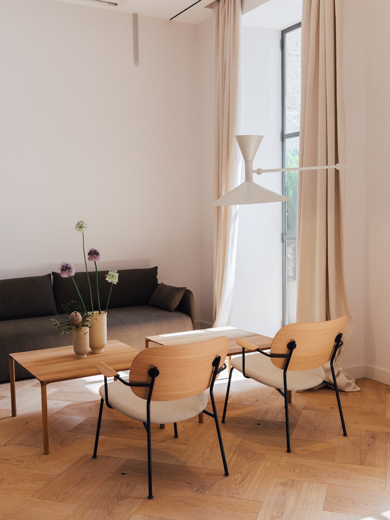 Minimal living area with oak parquet, two light wood chairs with black metal frames, a wooden coffee table, grey sofa, and