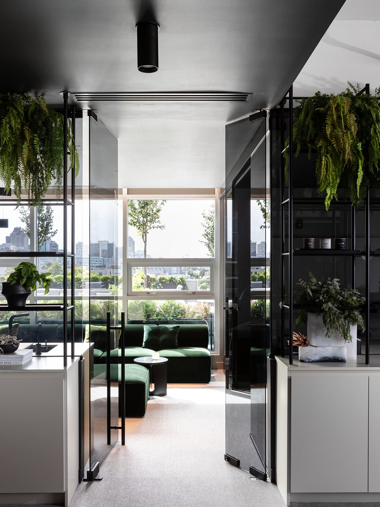 Modern office lounge with green velvet sectional, black glass partitions, hanging ferns and city view through large windows