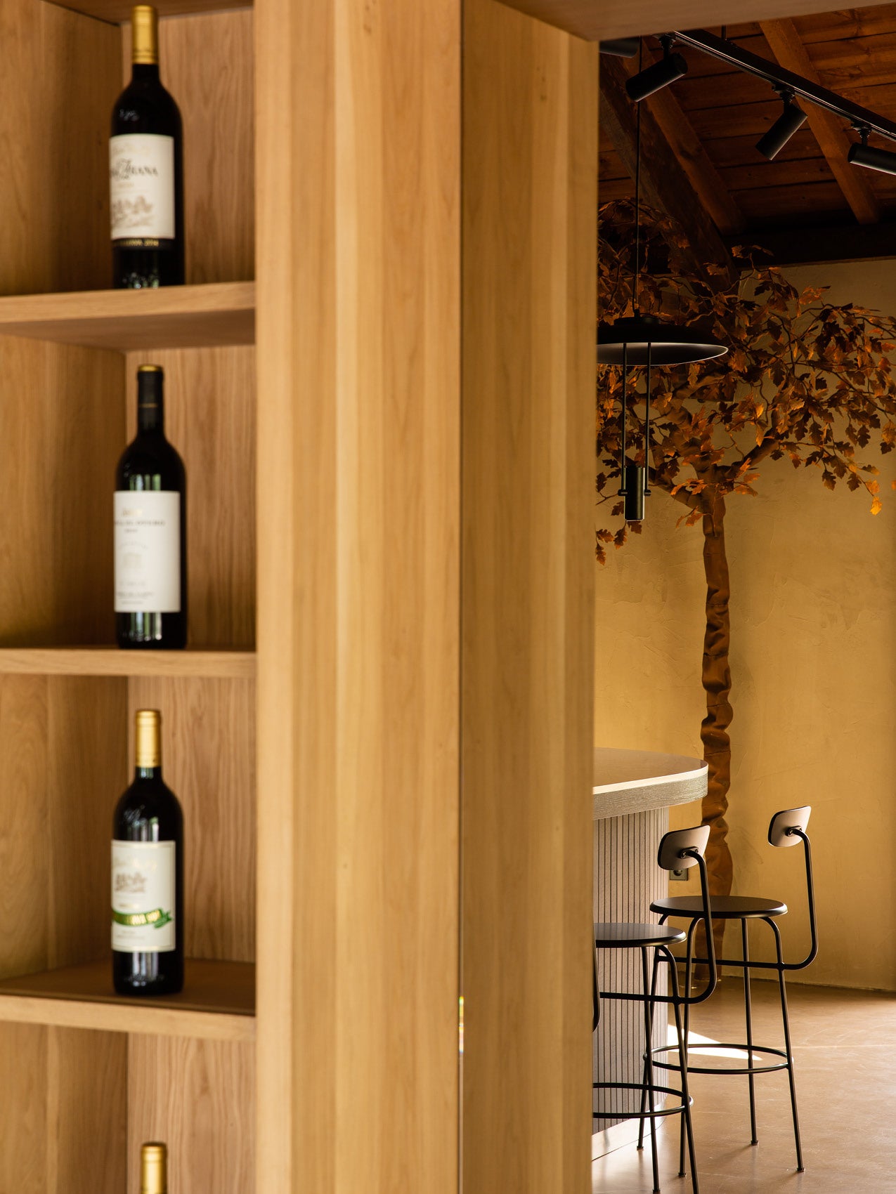 Oak shelving with three wine bottles visible, adjacent to a curved light-grey bar, black metal stools and hanging black