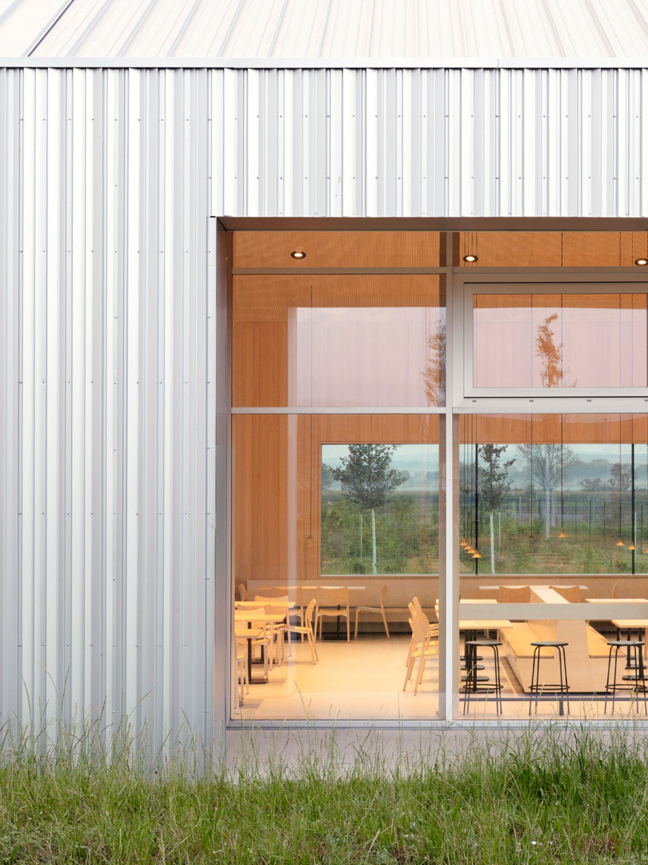 Exterior view of a corrugated white metal building with large glass window revealing warm wood interior and dining furniture